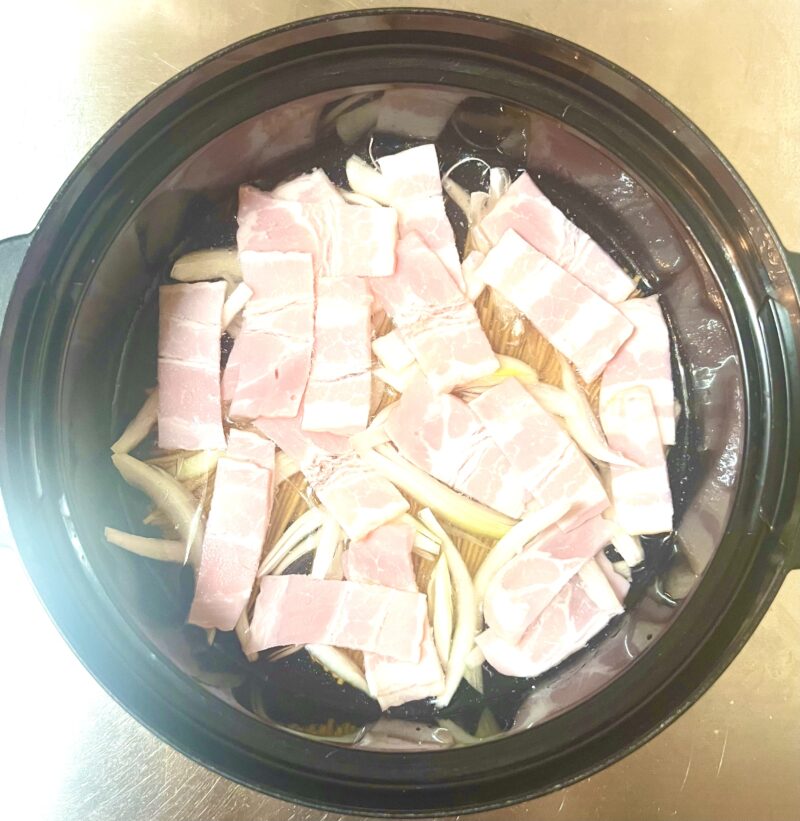 耐熱容器にに半分に折ったパスタ、薄切り玉ねぎ、1cm幅に切ったハーフベーコン、水、コンソメを入れた、レンジ加熱前の準備状態。